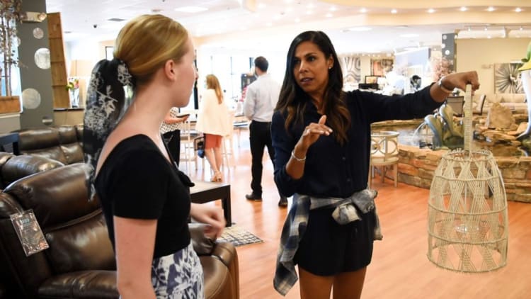 Two women in a modern furniture store; one holds a woven rattan pendant light shade with a handle, showcasing home decor options.