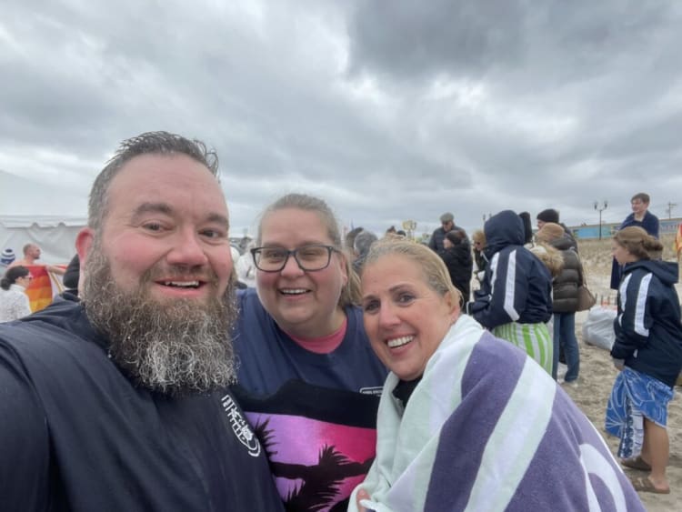 Three people standing close together outdoors on a cloudy day, one wrapped in a purple and white striped towel, with others in the background wearing jackets and beachwear, suggesting a cool beach event or gathering.