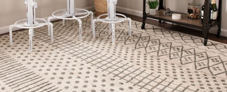 Beige and gray patterned area rug with geometric designs, placed under white metal bar stools and near a dark wood shelving unit.