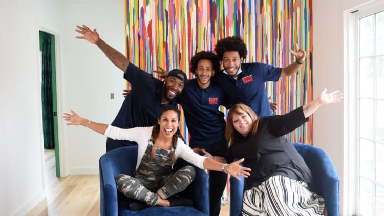 Five people posing energetically in a bright room with colorful vertical stripes on the wall, three wearing navy blue shirts with "BOB'S" logo.