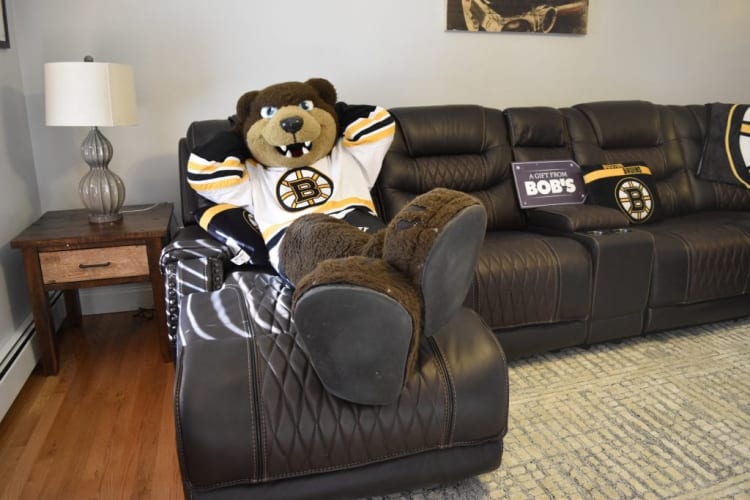 A plush Boston Bruins mascot in a white and yellow hockey jersey lounging on a dark brown leather sectional sofa next to a wooden side table with a glass lamp. The sofa has a sign that reads "A GIFT FROM BOB'S" and Bruins-themed pillows.