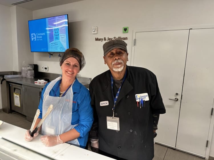 Two staff members at Covenant House New York wearing protective aprons and gloves, standing behind a counter in a kitchen or food service area. One holds tongs, the other wears a black chef coat with pens and a name tag. A screen in the background displays information about being a trans ally.