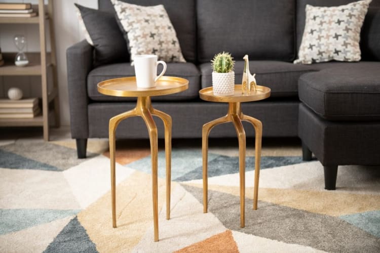 Two gold metal side tables with round tops on a geometric patterned rug in front of a dark gray sectional sofa with patterned pillows. One table holds a white ceramic mug, the other a small potted cactus and a gold giraffe figurine.