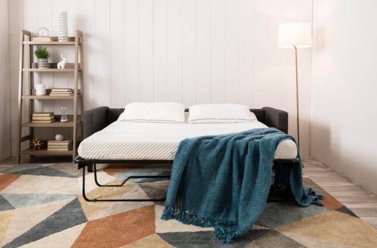 Modern living room with a dark gray sofa bed featuring white mattress and pillows, blue knitted throw blanket, wooden shelf with decor, and a floor lamp.