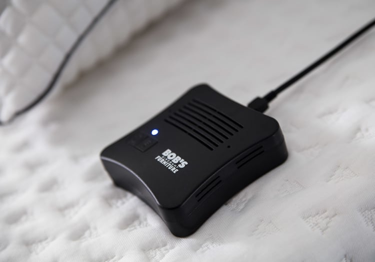 Black BOBIS furniture electronic device with ventilation slots and a blue indicator light, connected by a black cable on a textured white surface.