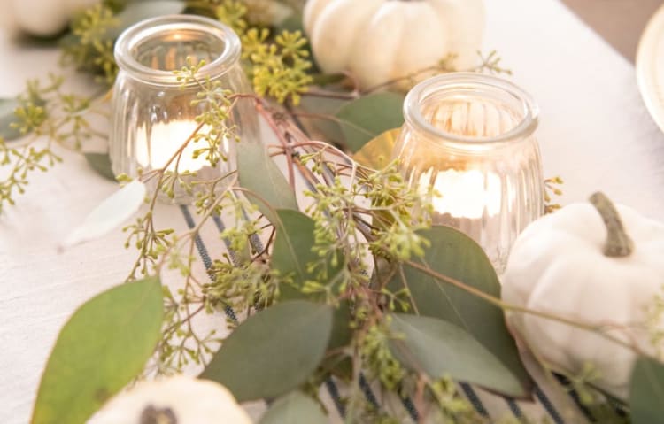 White mini pumpkins and eucalyptus greenery arranged with lit glass votive candles on a beige table runner, creating a cozy fall centerpiece.