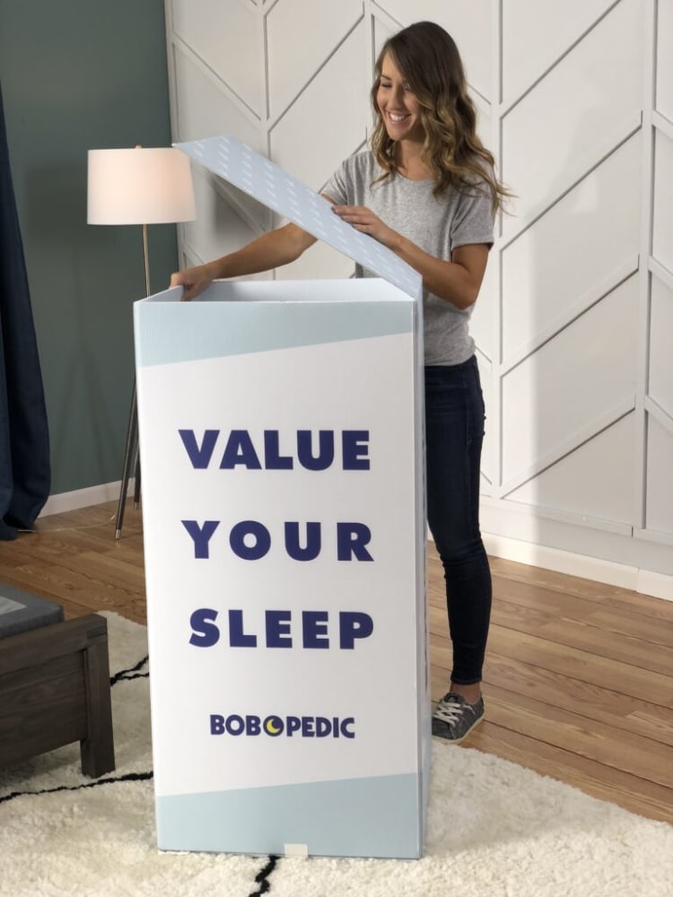 A person opening a tall rectangular box with text "VALUE YOUR SLEEP" and brand name "BOBOPEDIC" on the front, promoting sleep products.