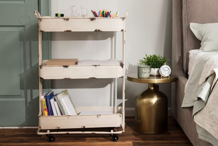 A vintage-style cream metal rolling cart with three shelves holding books, colored pencils, and a glass pitcher, next to a gold round bedside table with a green plant, a coffee mug with "but first COFFEE" text, and a silver alarm clock, beside a beige upholstered bed with layered bedding.