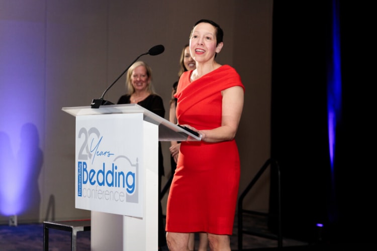 Woman in a red knee-length dress speaking at a podium with a sign reading "20 Years Furniture Today Bedding Conference" at an event.