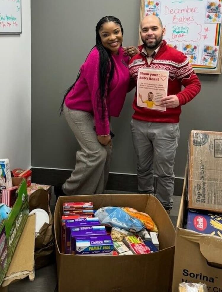 Two people stand in an office with boxes of assorted snacks including Cheez-It family size crackers. One holds a sign reading "Show your Bob's Heart" with a heart symbol and a cartoon man, promoting kindness and community support. A whiteboard behind them lists "December Babies" and colorful notes.