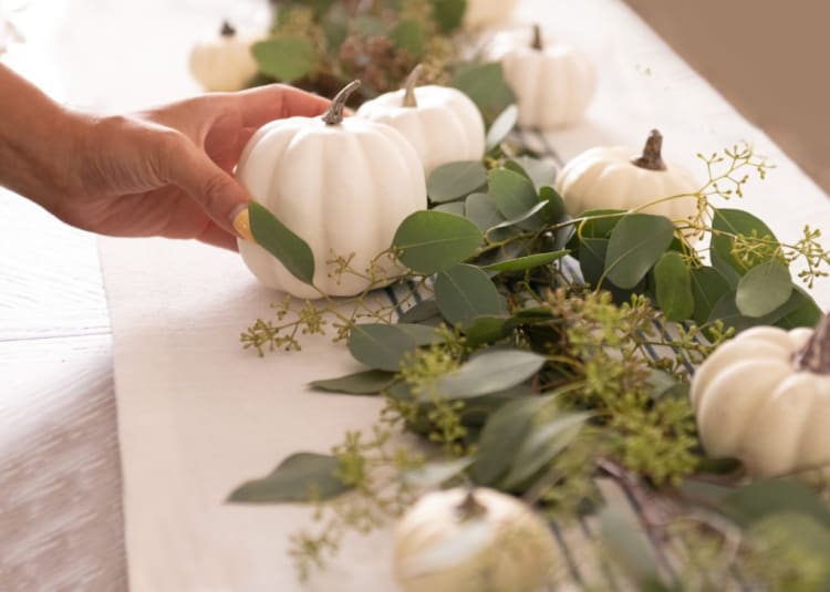 Hand placing a small white decorative pumpkin on a table runner adorned with green eucalyptus leaves, creating a natural fall centerpiece.