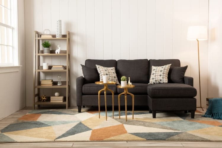 Dark gray fabric sectional sofa with patterned throw pillows, gold nesting side tables with decor, wooden ladder shelf, geometric area rug, and floor lamp in modern living room.