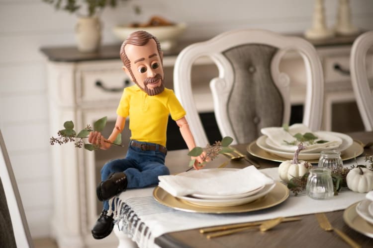 Articulated doll wearing a yellow shirt and blue jeans sitting on a dining table set with gold-rimmed plates, white napkins, small white pumpkins, and glass candle holders.
