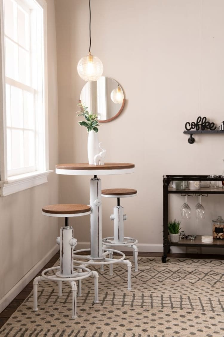 White and wood adjustable-height industrial-style bar table set with two matching stools, a white vase with green foliage, a hanging globe pendant light, round wall mirror, and a coffee-themed shelf in a bright, minimalist room with patterned rug.