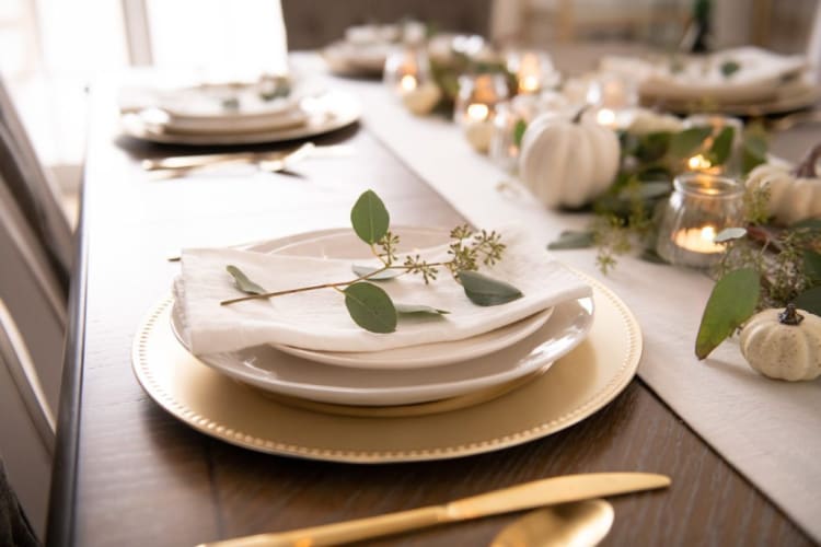 Elegant fall-themed table setting with white ceramic plates, gold charger, linen napkin, eucalyptus sprig, and small white pumpkins on a wooden table.