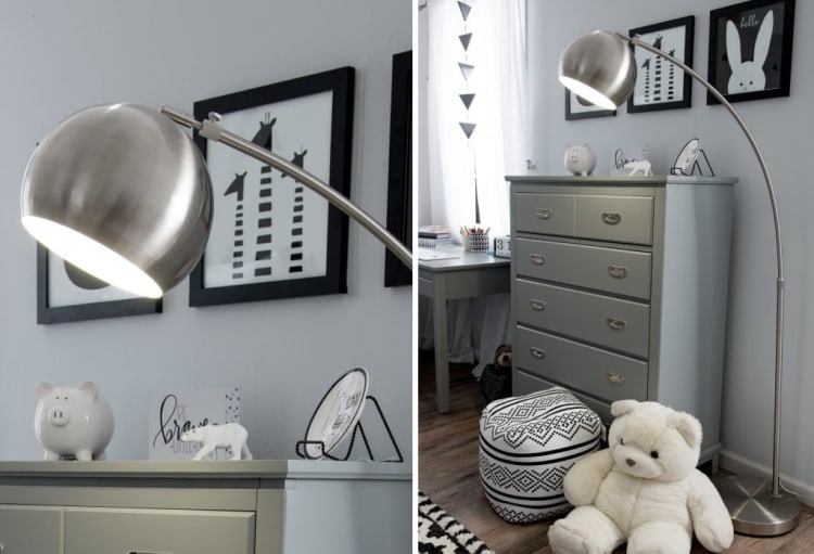 Modern brushed metal arc floor lamp with a large dome-shaped shade illuminating a gray chest of drawers in a minimalist children's room with black and white animal wall art, a white teddy bear, and patterned pouf on wood flooring.