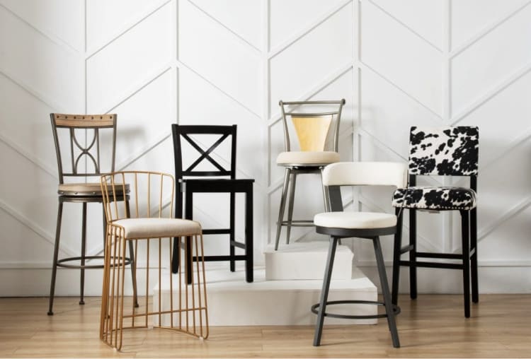 Six stylish bar stools with different designs and materials, including metal, wood, fabric, and cushioned seats, displayed on a wooden floor against a white geometric wall.