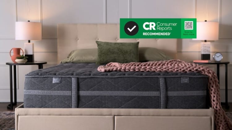 King-size mattress in dark gray fabric with quilted top and side handles, placed on beige upholstered bed frame with green pillows and pink knitted throw. Text overlay: "CR Consumer Reports RECOMMENDED Scan to learn more" with checkmark and QR code.