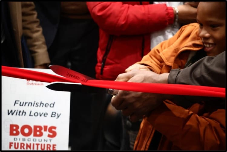 Hands cutting a red ribbon at a grand opening event with a sign reading "Furnished With Love By BOB'S Discount Furniture" in the background.