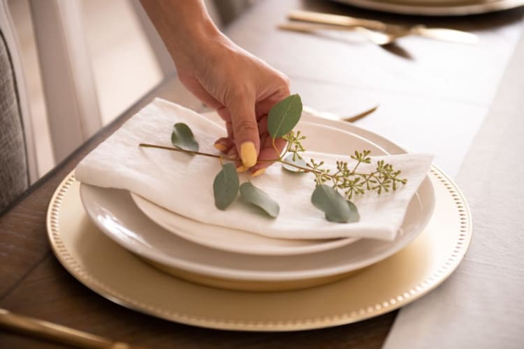 Hand placing a green eucalyptus sprig on a white cloth napkin, set on stacked beige and gold-rimmed ceramic plates on a wooden table.