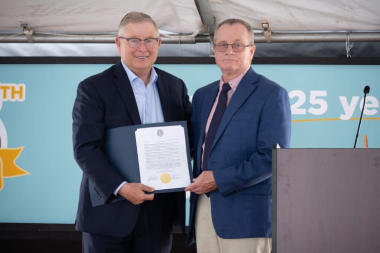 Two men in business attire stand side by side at a formal event, holding a certificate with official seals, against a blue backdrop with partial text "25 ye".