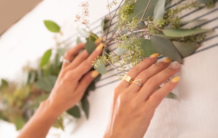Hands with yellow and white polka dot nail polish arranging green eucalyptus leaves on a striped fabric surface, showcasing natural decor and manicure details.