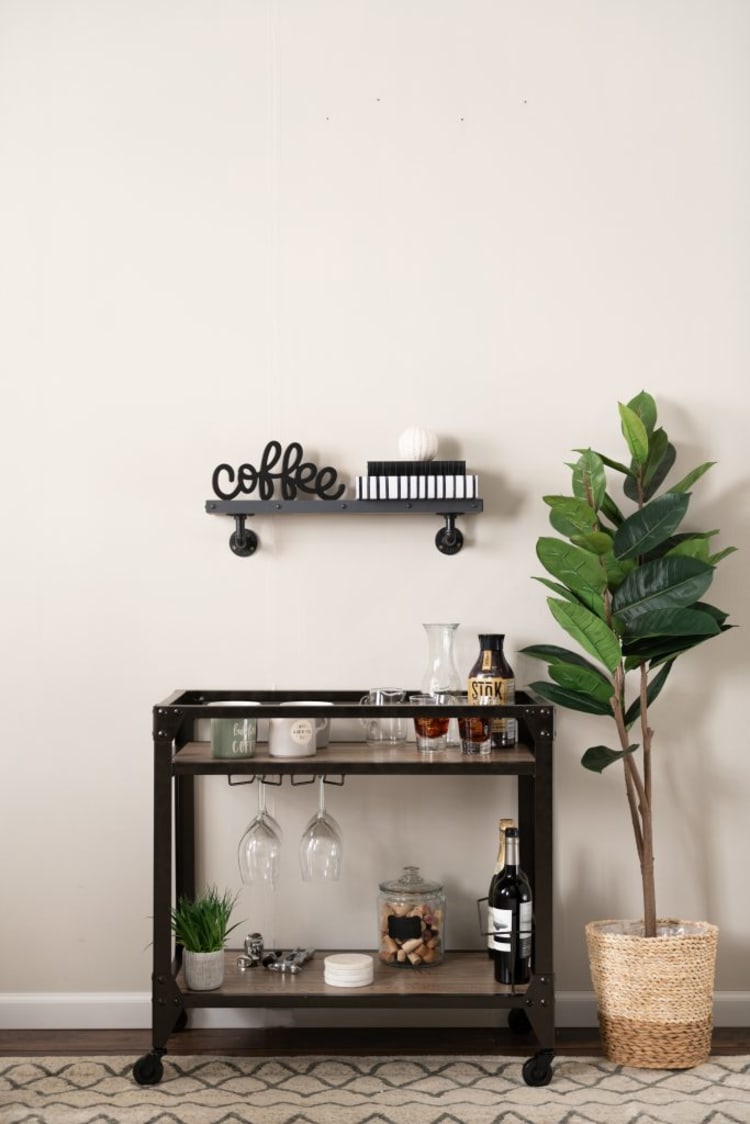 Black metal and wood coffee cart with mugs, wine bottles, glassware, and a potted plant, next to a tall green plant in a woven basket.