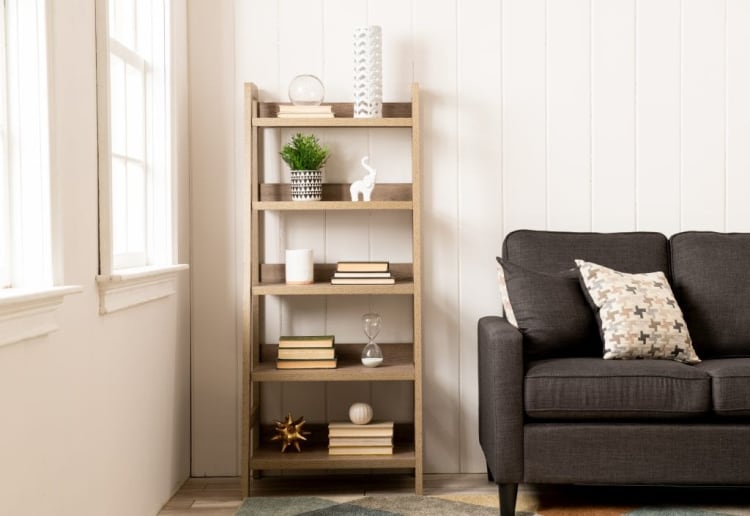 Wooden ladder-style bookshelf with decorative items and books next to a dark gray fabric sofa with patterned cushions in a bright living room.