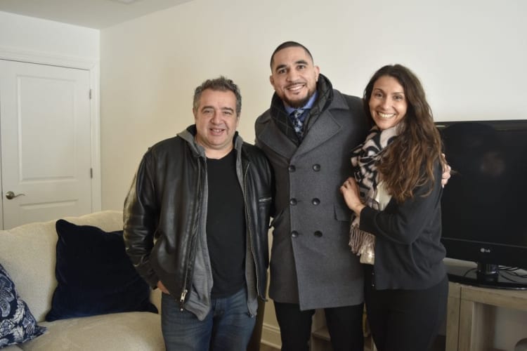 Three people standing indoors near a beige couch and an LG flat-screen TV, wearing casual and winter clothing including a black leather jacket, gray coat, and scarf.