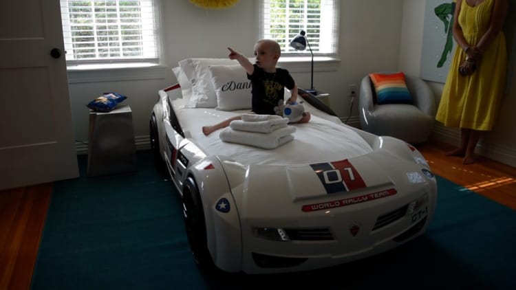 Child sitting on a white race car-themed bed with "01" and "World Rally Team" decals, in a bright room with blue carpet and yellow dress.