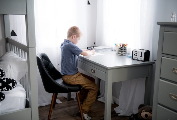 Young boy sitting at a gray wooden desk writing with a pencil, next to a black chair, a calendar showing "31 January," and a cup holding colorful pencils.