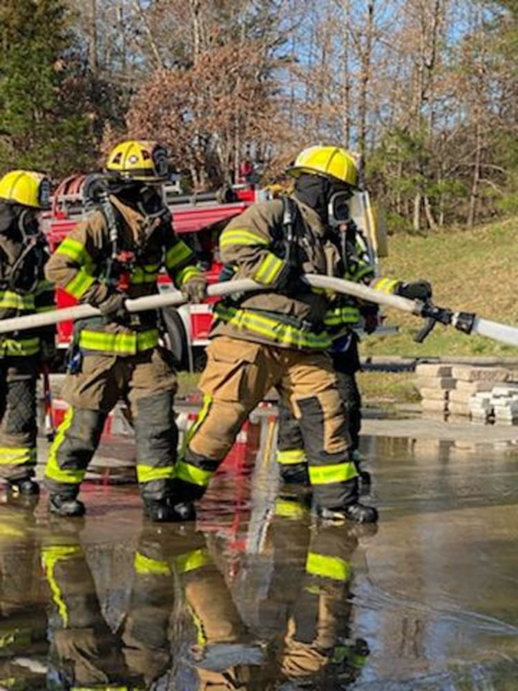 Firefighters in full protective gear and yellow helmets operate a large fire hose, spraying water during a training or emergency response.