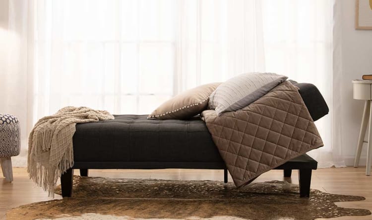 Dark gray upholstered chaise lounge with wooden legs, adorned with beige and gray throw pillows and a beige knitted blanket, set on a brown patterned rug in a bright room with sheer white curtains.