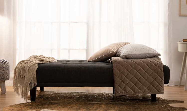 Dark gray upholstered bench with wooden legs, styled with beige throw blanket, quilted brown coverlet, and two decorative pillows in a bright, airy room.