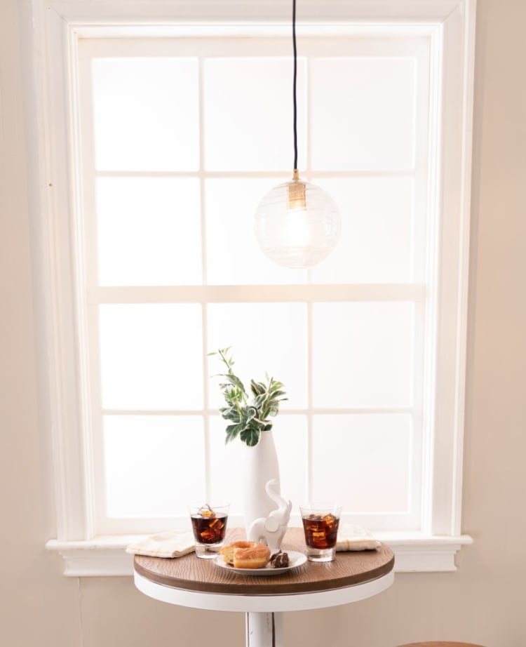 Round wooden table with two glasses of iced cola, a white vase with green foliage, a white ceramic elephant, and a plate with a donut and chocolate, set against a bright window with a hanging glass pendant light.