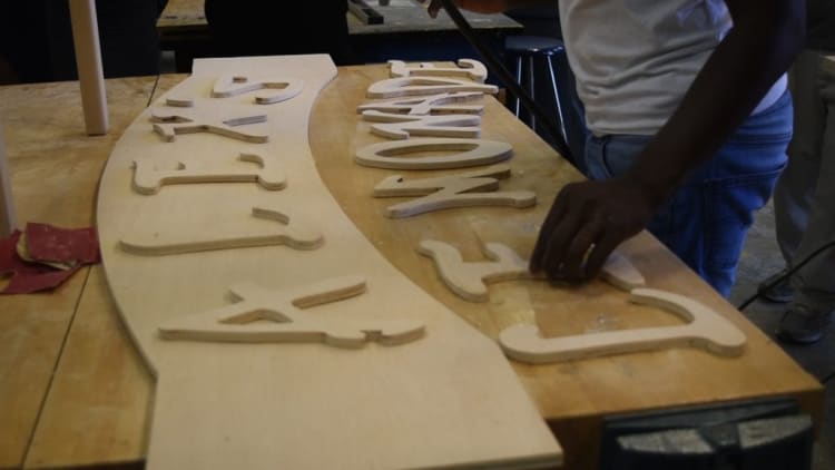 Wooden cut-out letters arranged on a workbench, spelling "FAMILY" and "EMPOWERMENT," with a person positioning the pieces, highlighting custom woodcraft signage.