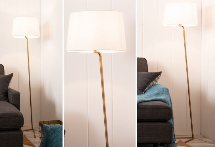 Modern floor lamp with a slim gold metal stand and white fabric drum shade, positioned next to a dark gray sofa with blue throw blanket.