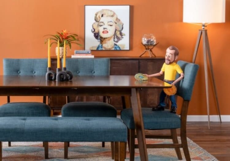 Modern dining room with teal upholstered chairs and bench, dark wood table, orange accent wall, decorative candles, plant, and tripod floor lamp.
