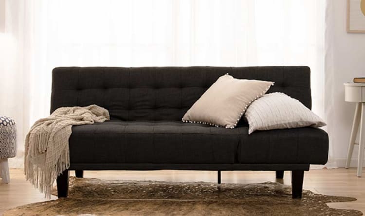 Dark gray tufted fabric sofa with wooden legs, adorned with beige and cream textured throw pillows and a fringed beige throw blanket, set in a bright living room with sheer white curtains and a brown patterned rug.