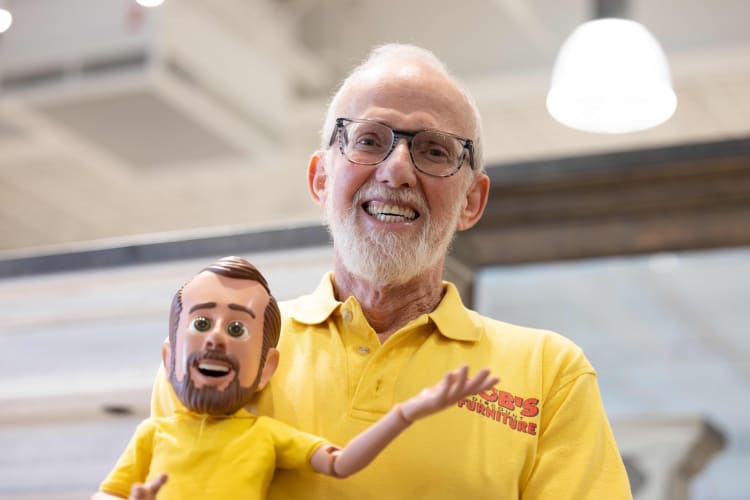 Older man wearing a yellow polo shirt with "Bob's Furniture" logo holding a yellow-shirted articulated doll in a bright indoor setting.