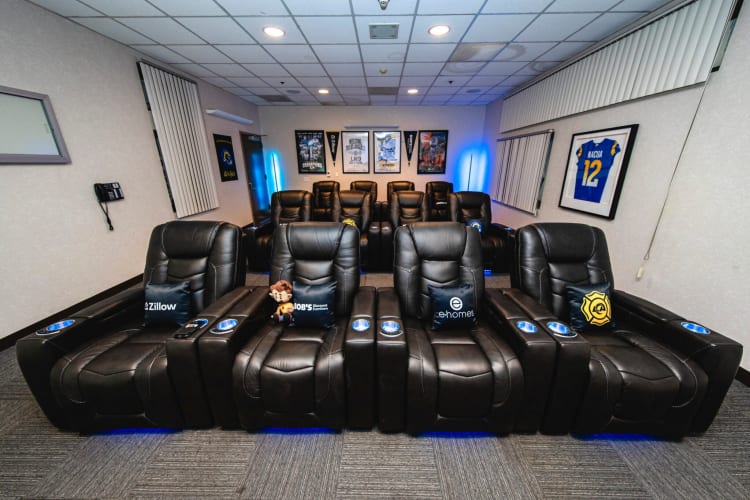 Black leather reclining theater chairs with branded pillows from Zillow, Bob's Discount Furniture, and e-homes, arranged in two rows with blue LED lighting in a modern home theater room.