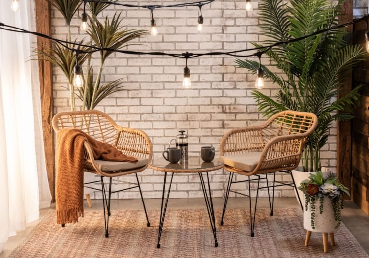 Cozy seating area with two rattan chairs featuring beige cushions, a round glass-top table, warm string lights, and green potted plants on a patterned rug.