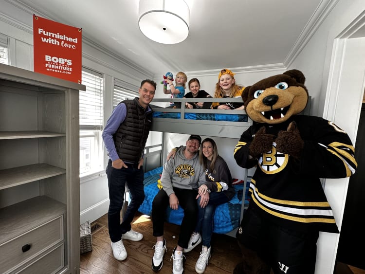 A family gathered in a bedroom with gray bunk beds and blue bedding, alongside a person in a Boston Bruins mascot costume. A red sign reads "Furnished with love BOB'S DISCOUNT FURNITURE MYBOBS.COM." The room features white walls, hardwood floors, and a gray wooden shelving unit.
