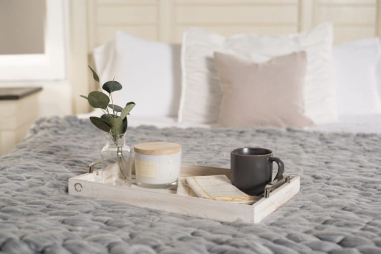 Wooden tray on a gray chunky knit blanket with a glass vase holding green eucalyptus, a white scented candle labeled "SEDG," and a black ceramic mug, styled on a bed with white and beige pillows.