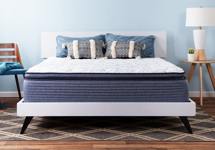 Modern bedroom setup featuring a white platform bed with a thick blue mattress, white quilted cover, decorative blue and patterned pillows, wooden side table with lamp and cactus, and a wooden chair with blue cushion against a light blue wall.
