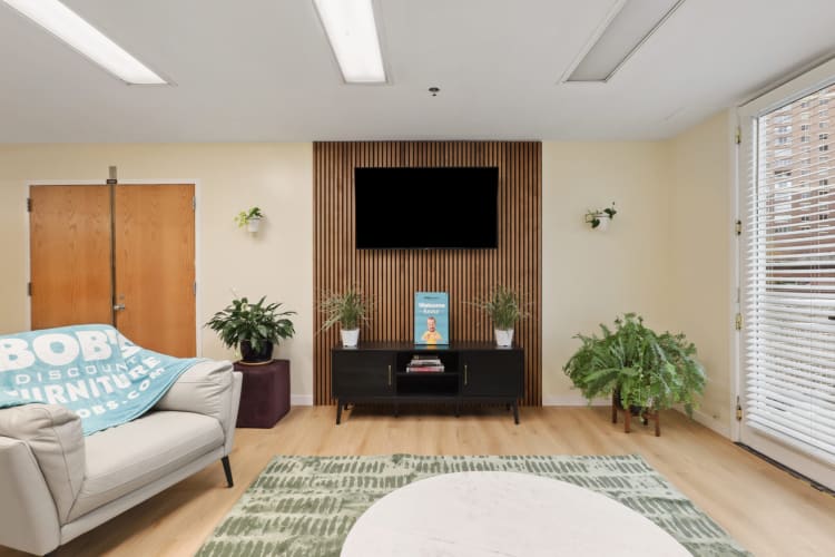 Modern living room with light wood flooring, beige walls, a black TV mounted on a vertical wood slat accent wall, black media console, green plants, and a beige armchair with a blue "BOB'S DISCOUNT FURNITURE" throw blanket.