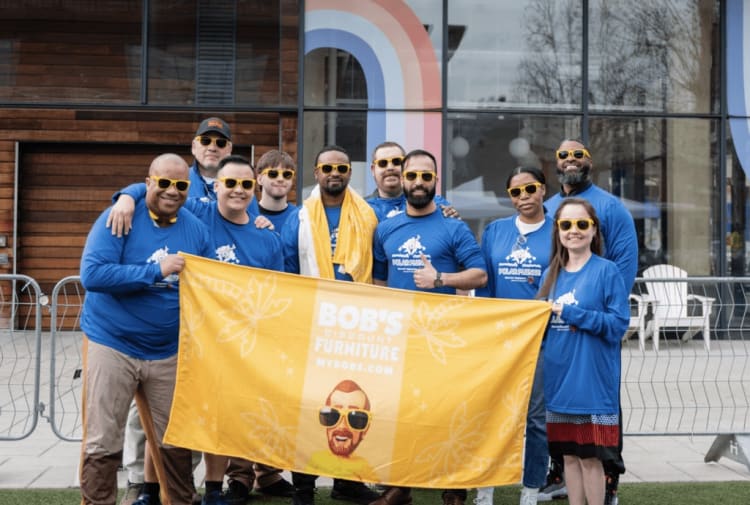 Group of people wearing blue Polar Plunge shirts holding a yellow banner that reads "BOB'S DISCOUNT FURNITURE MYBOBS.COM" in front of a modern building.