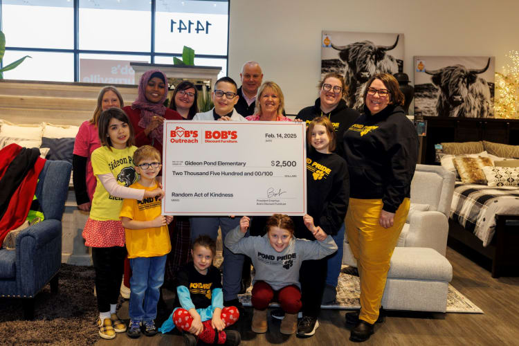 Group of adults and children holding a large check from Bob's Discount Furniture for $2,500 to Gideon Pond Elementary, dated Feb. 14, 2025, in a furniture store setting.