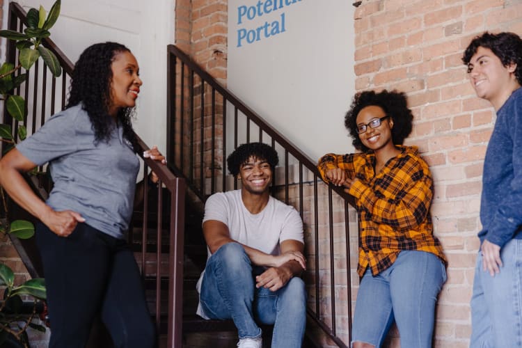 Four people casually gathered on and around a staircase with brick walls, featuring a woman in a yellow plaid shirt and jeans, a man in a white t-shirt and jeans sitting on the stairs, a woman in a gray t-shirt and black pants standing, and a man in a blue sweater and jeans standing near a wall. Text on the wall reads "Potential Portal."