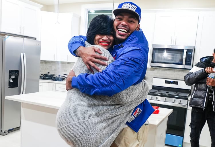 Two people embracing in a modern white kitchen with stainless steel appliances, including a refrigerator, microwave, and stove. One person wears a blue jacket and a blue Giants cap, while the other wears a gray knit sweater. Another person in a black and white jacket holds a water bottle in the background.